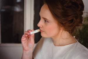 Woman in a white dress applying cocoa butter lip therapy chapstick, a natural lip balm perfect as the best anti aging lip balm moisturizer for chapped lips.