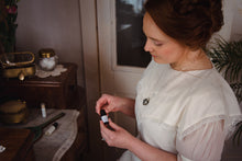 Woman in white Victorian dress wearing antique-style aromatherapy scent locket perfume locket by Willow & Birch Apothecary and holding essential oil blend while sitting at antique marble-topped vanity table