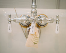 Close-up of a shiny bathroom tap with 'HOT' and 'COLD' labels, adorned with a small linen bag of Willow Birch Apothecary spa bath salts, emphasizing the soothing epson salt and therapeutic salts for a spa-like experience at home.