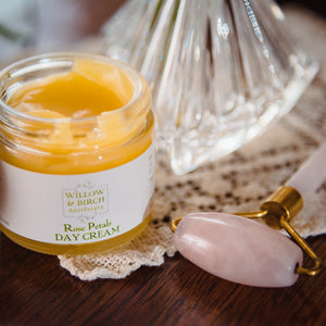 Open jar of Rose Petals Day Cream by Willow & Birch Apothecary on a wooden surface with a rose quartz facial roller, symbolizing the best face moisturizer for dry face and a good cream for wrinkles.