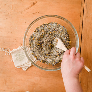 A person's hand mixing dried herbs into a bowl of natural bath salts, creating a blend of therapeutic salts. This image captures the preparation of spa bath salts, highlighting the epsom salt uses and benefit of epsom salt bath.