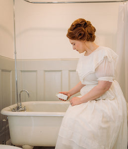 Woman in Victorian dress pouring bath salts by Willow & Birch Apothecary into antique clawfoot bath tub